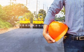65814842-engineer-holding-safety-helmet-at-road-construction-site-with-roller-compactor-working-asphalt-road